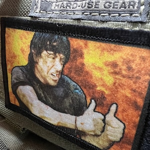 May include: A black and white patch with a person giving a thumbs up. The background is a fiery orange and yellow. The patch has the text "Hard-Use Gear" on it.