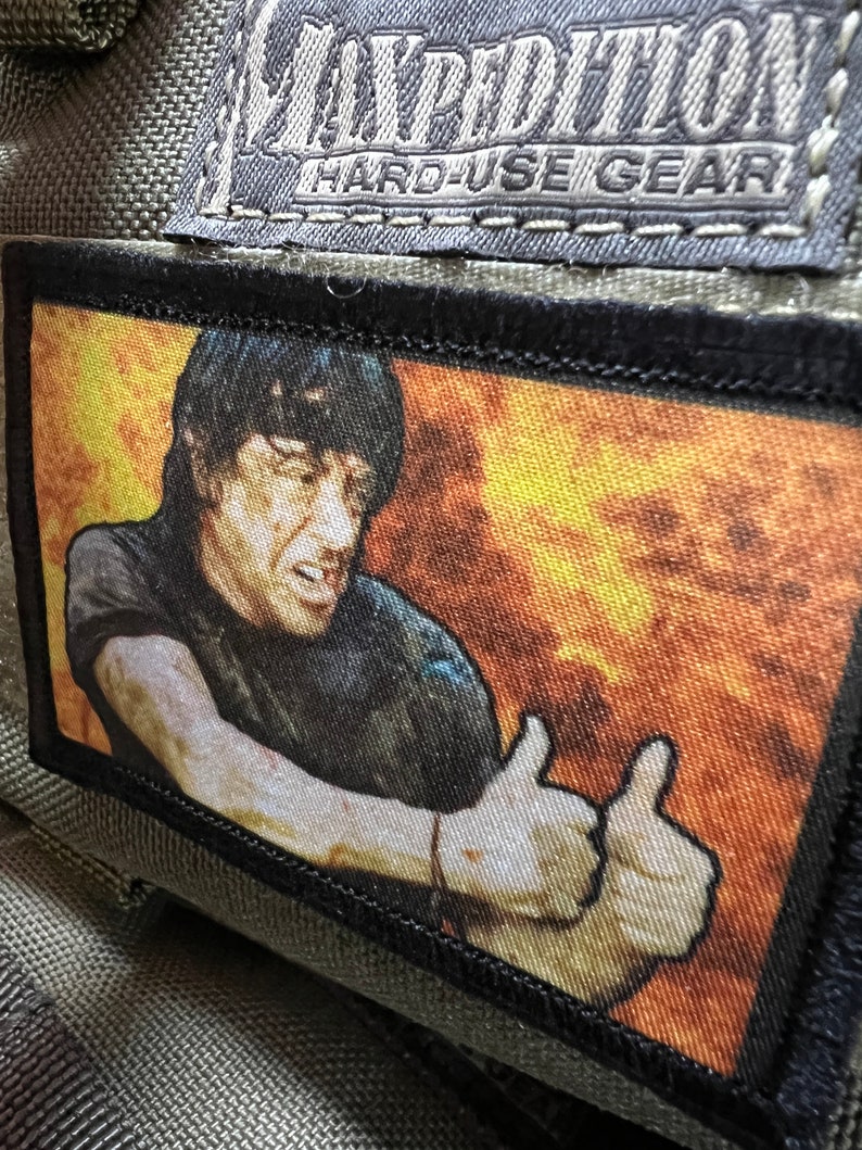 May include: A black and white patch with a person giving a thumbs up. The background is a fiery orange and yellow. The patch has the text "Hard-Use Gear" on it.
