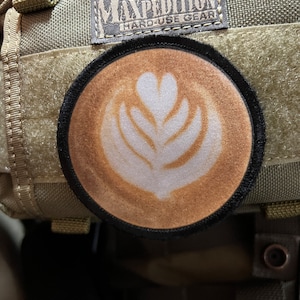 Peut inclure: Un patch brun et noir avec un motif de café blanc sur un sac à dos camouflage vert et brun. Le patch porte le texte "Maxpedition Hard-Use Gear".