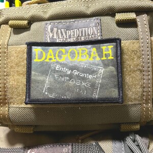 May include: A rectangular morale patch with a black border featuring the yellow text DAGOBAH above a graphic of a passport stamp that reads Entry Granted. The patch is attached to a tan Maxpedition gear bag with a hook and loop fastener system.