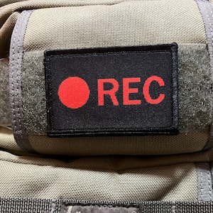 Könnte beinhalten: Ein schwarzer Patch mit einem roten Kreis und dem Wort "REC" in weißen Buchstaben. Der Patch ist an einer beigefarbenen Stofftasche befestigt.