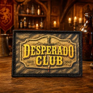 May include: A rectangular embroidered patch with a black border featuring the text DESPERADO CLUB in bold yellow letters. The design includes a central sword graphic set against a textured, weathered background with ornate decorative elements.