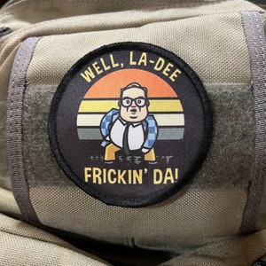 Könnte beinhalten: Ein gestickter schwarzer und gelber Aufnäher mit einer Comicfigur, die ein blau-weiß kariertes Hemd und eine Brille trägt. Der Aufnäher trägt den Text "Well, La-Dee Frick'n' Da!" in gelber Schrift.