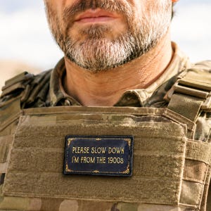May include: A rectangular black patch with gold decorative borders and gold text that reads, PLEASE SLOW DOWN I'M FROM THE 1900s. The patch is attached to the front of a camouflage tactical vest with a hook-and-loop fastener surface.