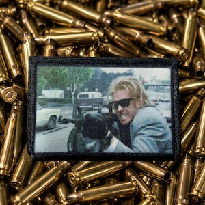 May include: A rectangular fabric patch with a black border featuring a printed scene of a person with blonde hair wearing sunglasses and holding a rifle, set against a background of numerous scattered brass-colored metallic bullet casings.