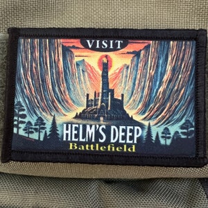 Op de afbeelding: Een rechthoekige patch met een zwarte rand met een kleurrijke illustratie van een fort bij zonsondergang. De tekst "VISIT HELM'S DEEP Battlefield" staat boven en onder de afbeelding.