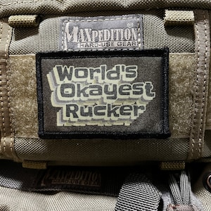 以下が含まれることがあります： 「World's Okayest Rucker」と書かれた黒いパッチと、「Maxpedition Hard-Use Gear」と書かれた白黒のラベルが付いた緑色のミリタリースタイルのバックパック。