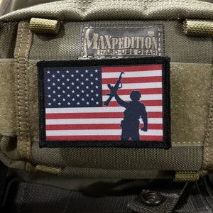 Pode incluir: Uma mochila tática verde com um remendo preto que mostra uma bandeira americana e a silhueta de uma pessoa segurando um rifle. O remendo tem o texto "Maxpedition Hard Use Gear" acima da bandeira.
