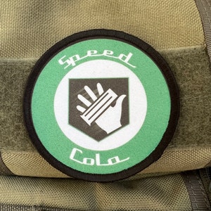 Puede incluir: Un parche redondo verde y blanco con el texto "Speed Cola" y una mano estilizada sosteniendo una lata. El parche tiene un borde negro.