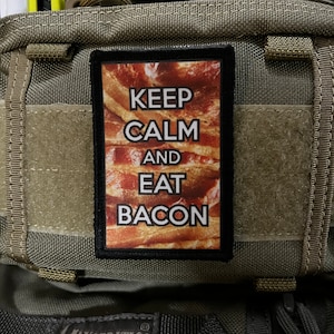 May include: A green tactical backpack with a black patch that reads "Keep Calm and Eat Bacon". The patch features a background image of bacon.