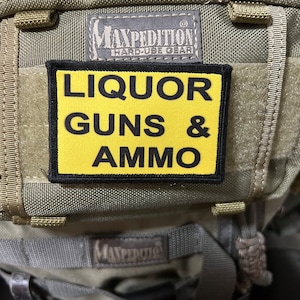 May include: A tactical backpack with a yellow patch that reads "LIQUOR GUNS & AMMO" in black block letters. The patch is rectangular and is attached to the front of the backpack. The backpack is olive green and has multiple straps and pockets.