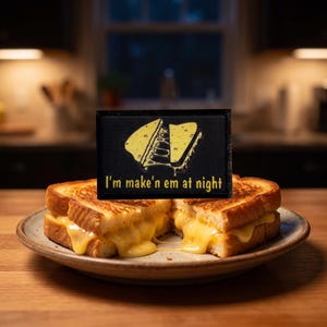 Puede incluir: Un parche rectangular negro con un gráfico amarillo de un sándwich de queso a la parrilla con hilos de queso derretido y el texto "I'm make'n em at night" en letras amarillas, colocado frente a un sándwich de queso en un plato.