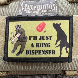 Op de afbeelding: Een zwart en groene patch met een cartoon afbeelding van een persoon die een rode bal naar een hond gooit. De tekst op de patch luidt "I'm just a Kong dispenser."