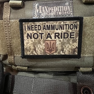 May include: A green military-style backpack with a black patch that reads "I NEED AMMUNITION NOT A RIDE" with a Ukrainian trident symbol. The backpack has a black label that reads "MAXPEDITION HARD-USE GEAR".