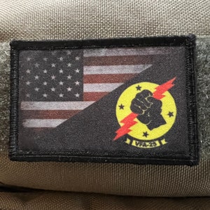 May include: A black patch with a yellow and red lightning bolt and a fist. The patch also has a small American flag in the top left corner. The text "VALCES" is visible on the patch.
