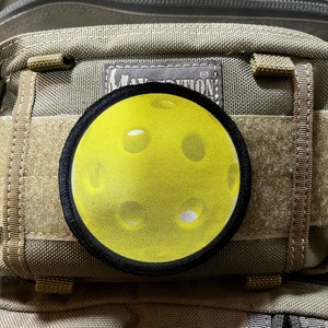 Puede incluir: Un parche amarillo y negro con un objeto redondo amarillo texturizado en el centro. El parche está unido a una bolsa marrón texturizada.