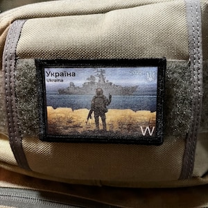 Pode incluir: Um remendo preto e branco com um soldado em pé numa praia. O remendo tem o texto "Україна" e "Ukraine" em cirílico e inglês, e o ano "2022".