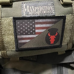 Op de afbeelding: Een groene militaire rugzak met een zwarte patch met een Amerikaanse vlag en een rood stierenlogo. De patch is op de voorkant van de tas genaaid. De tas heeft de tekst "Maxpedition Hard-Use Gear" bovenaan.