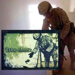 May include: A rectangular morale patch featuring a green and black graphic of a soldier in camouflage gear reaching down toward an object. The text Vibe check is printed in a light green font on the left side of the patch with a dark border.