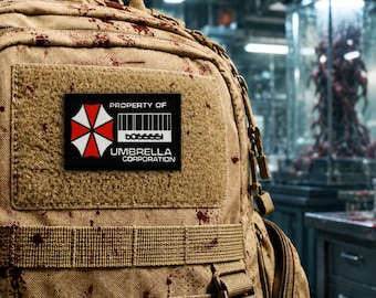 May include: A tan tactical backpack featuring dark red blood-like splatters across the fabric. A rectangular black patch is attached to the front, displaying a red and white umbrella logo, a barcode, and the text PROPERTY OF UMBRELLA CORPORATION.