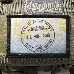 Op de afbeelding: Een groene militaire tas met een zwarte patch waarop "Ben Gurion Border Control" staat en een stempel met de tekst "Exit 12-09-2010". De tas heeft een zwart label met de tekst "Maxpedition Hard-Use Gear".