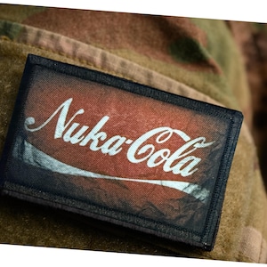 Peut inclure: Un écusson rectangulaire avec le texte "Nuka-Cola" en écriture blanche sur un fond rouge et orange. L'écusson a une bordure noire et est posé sur un tissu de camouflage.
