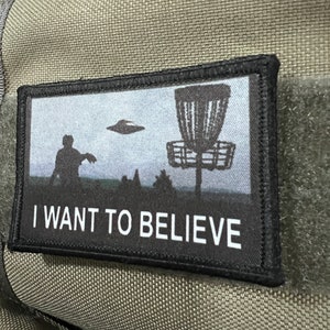 May include: A black and white patch with a disc golf basket and a silhouette of a person throwing a disc. The text "I WANT TO BELIEVE" is printed below the image.