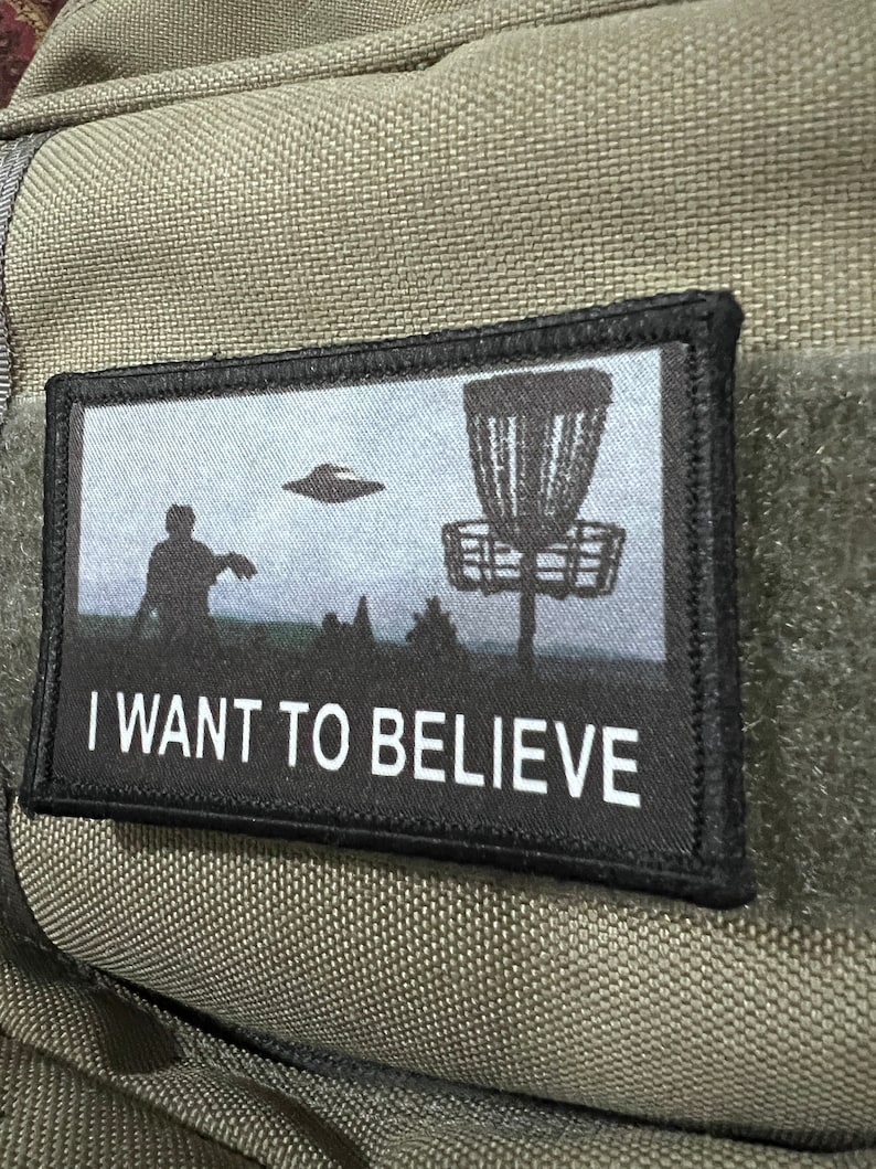 May include: A black and white patch with a disc golf basket and a silhouette of a person throwing a disc. The text "I WANT TO BELIEVE" is printed below the image.