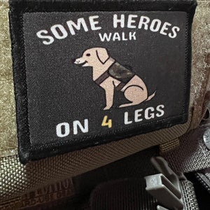Könnte beinhalten: Ein schwarzer Aufnäher mit dem Text "SOME HEROES WALK ON 4 LEGS" und einer Hundeillustration. Der Aufnäher ist auf einem olivgrünen Stoff angebracht, darüber das Logo "MAXPEDITION HARD-USE GEAR".