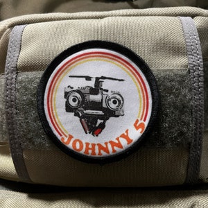 May include: A black and white patch with a red, orange, and yellow striped circle around it. The patch has a black and white image of a helicopter inside the circle. The text "JOHNNY'S" is written in black letters on the bottom of the patch.