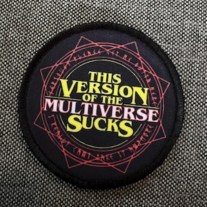 Puede incluir: Un parche redondo negro con un diseño circular rojo y blanco. El texto "This Version of the Multiverse Sucks" está escrito en letras amarillas.