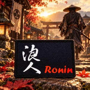 May include: A rectangular black patch with a stitched border featuring the white Japanese kanji for Ronin and the red text Ronin printed on the front. The patch is positioned in the foreground against a blurred background of a traditional setting.