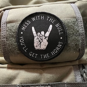 Może przedstawiać: Czarno-biała łatka z tekstem "Mess with the bull, you'll get the horns" i ręką wykonującą znak rock and roll.