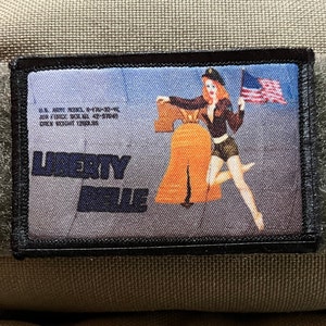May include: A rectangular patch with a pin-up girl illustration holding an American flag and standing next to the Liberty Bell. The patch has the words "LIBERTY BELLE" and military text. The patch is attached to a tan fabric.
