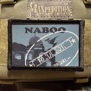 May include: A black and blue embroidered patch with the word "NABOO" and a stamp that says "10 JUL 2017" and "ARRIVAL..." The patch is attached to a green fabric background.