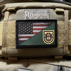 May include: A green, black, and yellow patch with an American flag and a military insignia. The patch is attached to a brown backpack with the text "Maxpedition Hard-Use Gear".