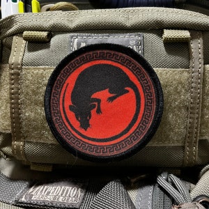 May include: A black and red embroidered patch with a stylized rat design inside a circle with a Greek key border. The patch is attached to a green tactical backpack.