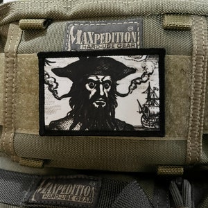 Op de afbeelding: Een groene Maxpedition Hard-Use Gear tas met een zwart-witte piratenpatch met een schedel en gekruiste beenderen. De patch is op de voorkant van de tas genaaid.