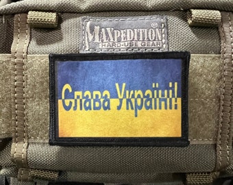 Puede incluir: Una mochila táctica verde con un parche negro que muestra la bandera de Ucrania y el texto "Слава Україні!" (Gloria a Ucrania!). El parche está unido a la parte delantera de la mochila.