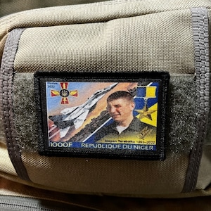 May include: A black and white patch with a blue and yellow Ukrainian flag, a jet fighter, and a portrait of a man. The text on the patch reads "Serpon Tarabaika 1993-2022" and "1000F Republique du Niger".