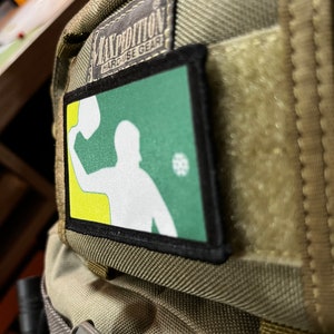 Pu&ograve; includere: Una toppa verde e gialla con una silhouette bianca di una persona che lancia una palla. La toppa &egrave; attaccata a uno zaino marrone con il testo "EXPEDITION HARDUSE GEAR" su un'etichetta nera.