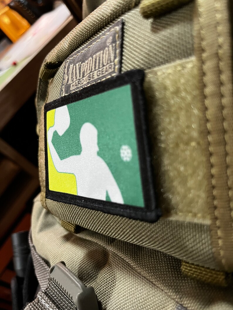 Pu&ograve; includere: Una toppa verde e gialla con una silhouette bianca di una persona che lancia una palla. La toppa &egrave; attaccata a uno zaino marrone con il testo "EXPEDITION HARDUSE GEAR" su un'etichetta nera.