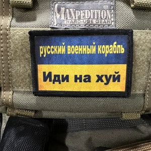 Puede incluir: Una mochila de estilo militar verde con un parche negro que dice "русский военный корабль Иди на хуй" en escritura cirílica. El parche está unido a la parte delantera de la mochila.