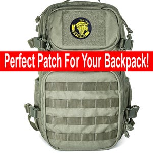 May include: An olive green tactical rucksack with multiple compartments and straps. A circular patch with the text "ZIP TIE CERTIFIED" and a parachute design is attached to the front. The text "Perfect Patch For Your Backpack!" is displayed in a red banner.