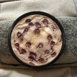 May include: A round patch featuring a detailed image of a chocolate chip cookie. The cookie has a light brown base with numerous dark brown chocolate chips. The patch is bordered by a black edge and is set against a tan and green background.
