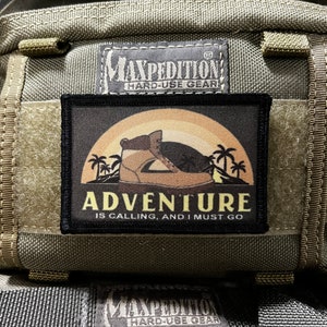Op de afbeelding: Een zwart en bruin patch met een zonsondergangscène en een laars met de tekst "Adventure is calling, and I must go" en het Maxpedition-logo met de tekst "Hard-Use Gear".