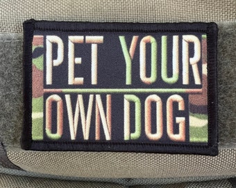 May include: A rectangular patch with the text "PET YOUR OWN DOG" in white and green lettering. The background features a camouflage pattern in shades of brown, green, and tan. The patch has a black border.