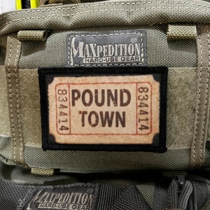 May include: A green Maxpedition bag with a black patch that says "Pound Town" and the numbers "834414" on either side. The bag has a black strap and a black zipper.