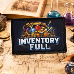 May include: A rectangular black patch featuring a colourful embroidered illustration of a treasure chest overflowing with gold coins, a red gemstone, a blue potion bottle, and a sword, with the white text INVENTORY FULL printed below.