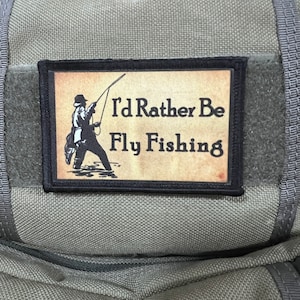 May include: A black and tan patch with a black border and the text "I'd Rather Be Fly Fishing" and a silhouette of a person fly fishing.